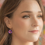 A woman with long brown hair and Vintage Burgundy Garden Pansy Drop Earrings from Holly Yashi Jewelry smiles softly, her makeup accentuating her eyes and complexion against a colorful, blurred background.