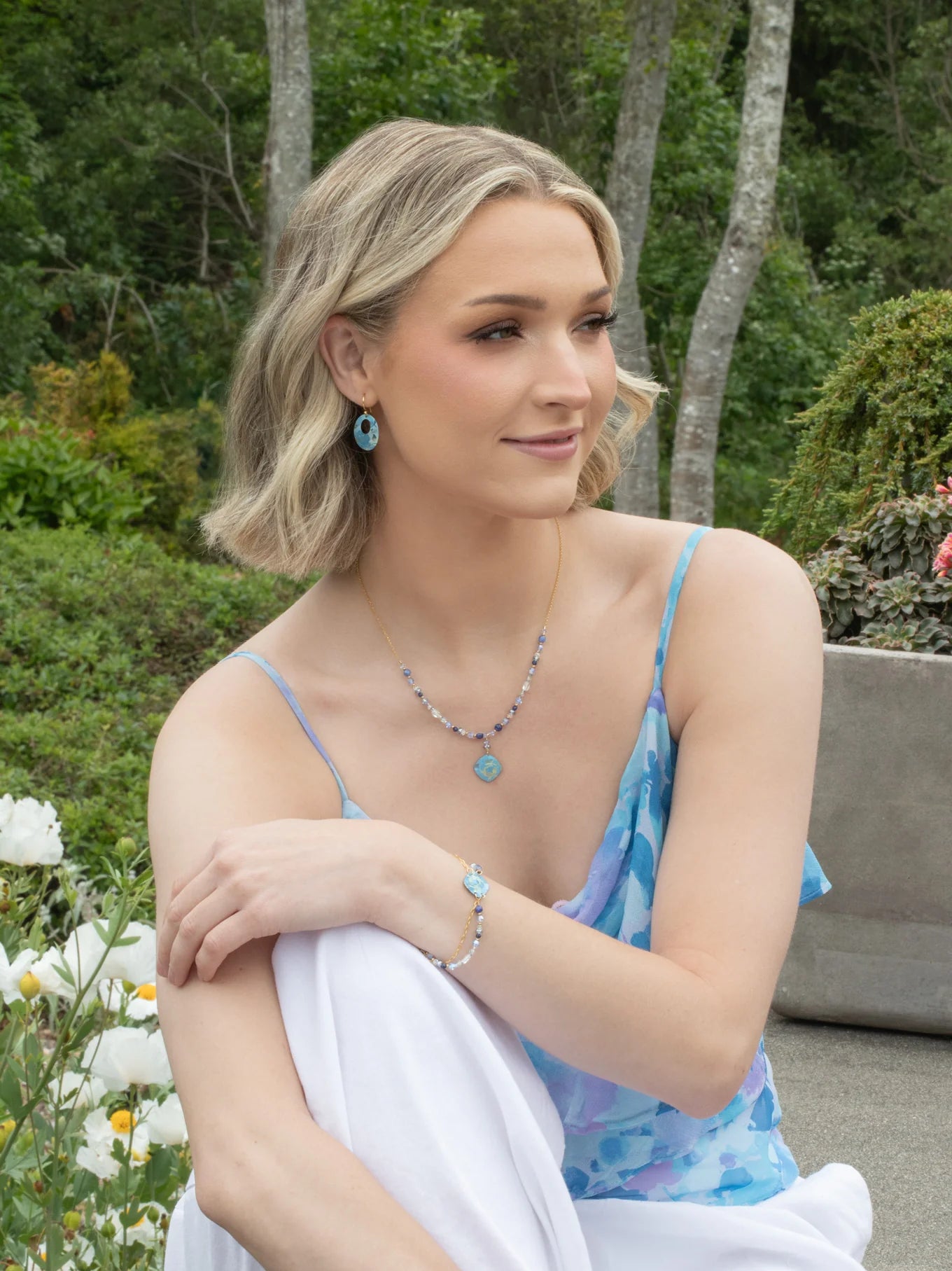 A woman with short blonde hair sits outdoors among greenery, wearing a blue floral dress and vintage-inspired jewelry, including matching blue earrings, a necklace, and the Elena Beaded Bracelet Sky Blue by Holly Yashi. She looks relaxed and gentle.