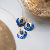 The Arabella Sapphire Blue Earrings by Holly Yashi feature bright blue, patterned pendants and dark blue gemstones, complemented by delicate glass beads and displayed near textured gold accents.