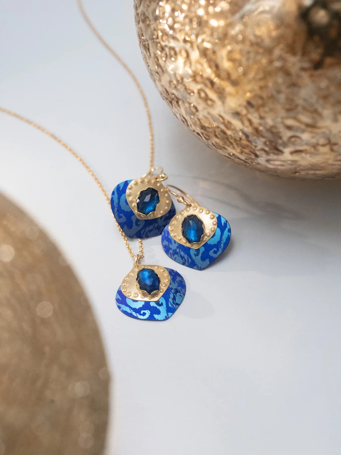 The Arabella Sapphire Blue Earrings by Holly Yashi feature bright blue, patterned pendants and dark blue gemstones, complemented by delicate glass beads and displayed near textured gold accents.