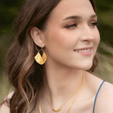 A woman with long brown hair smiles while looking to the side, adorned in Gold Ariel Earrings by Holly Yashi and layered gold-plated necklaces. Her blue outfit complements the blurred greenery in the background.