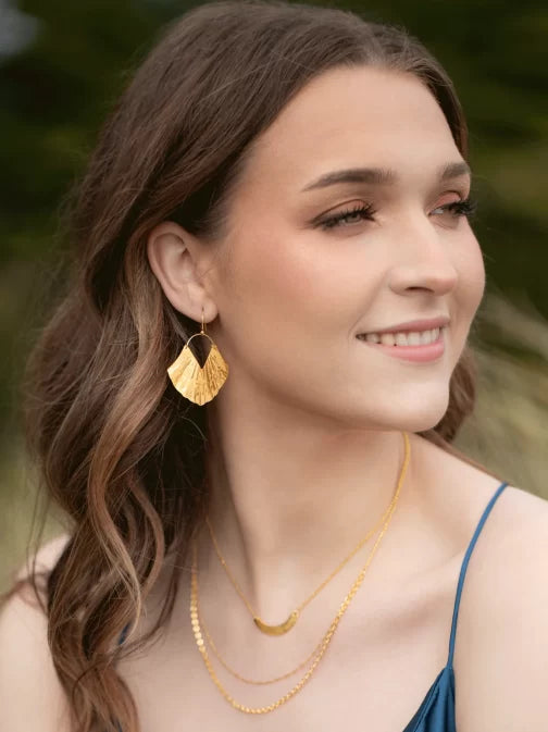A woman with long brown hair smiles while looking to the side, adorned in Gold Ariel Earrings by Holly Yashi and layered gold-plated necklaces. Her blue outfit complements the blurred greenery in the background.