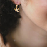 Close-up of a womans ear wearing Holly Yashis Plumeria Drop Gold Earrings. The blurred background highlights her curly hair and smooth skin, showcasing the intricate detail of this feminine jewelry from Holly Yashi.