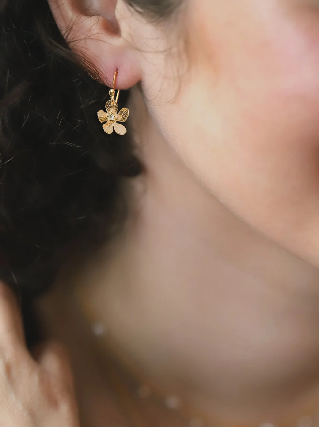 Close-up of a womans ear wearing Holly Yashis Plumeria Drop Gold Earrings. The blurred background highlights her curly hair and smooth skin, showcasing the intricate detail of this feminine jewelry from Holly Yashi.
