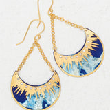 The Selena/ Midnight Blue earrings by Holly Yashi feature gold and blue crescent shapes with sunburst designs, small clear gemstones, and dangling gold chain hooks. Hypoallergenic and shown on a textured white surface.