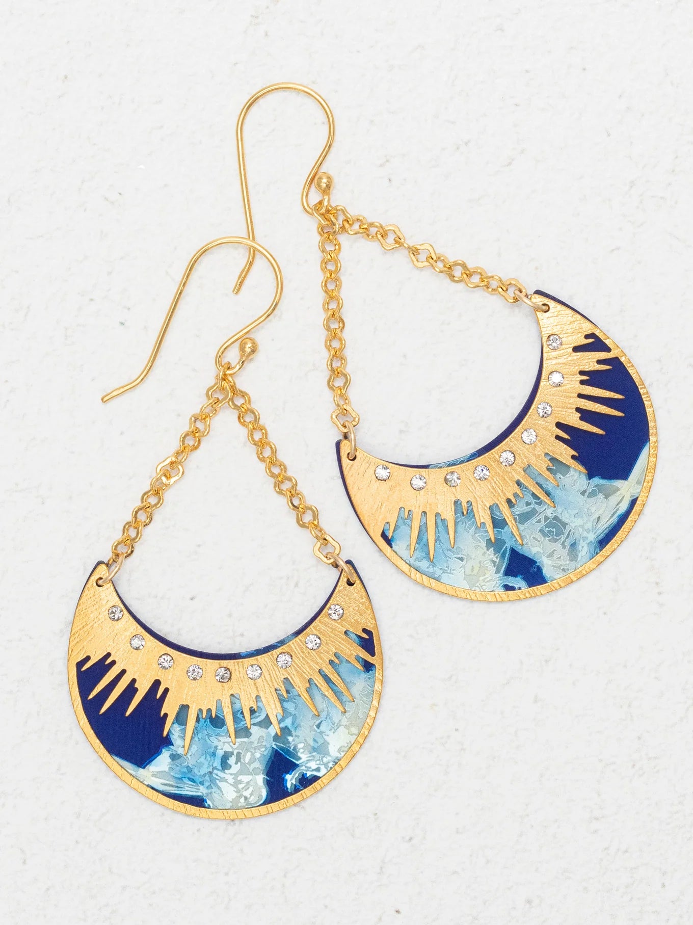The Selena/ Midnight Blue earrings by Holly Yashi feature gold and blue crescent shapes with sunburst designs, small clear gemstones, and dangling gold chain hooks. Hypoallergenic and shown on a textured white surface.