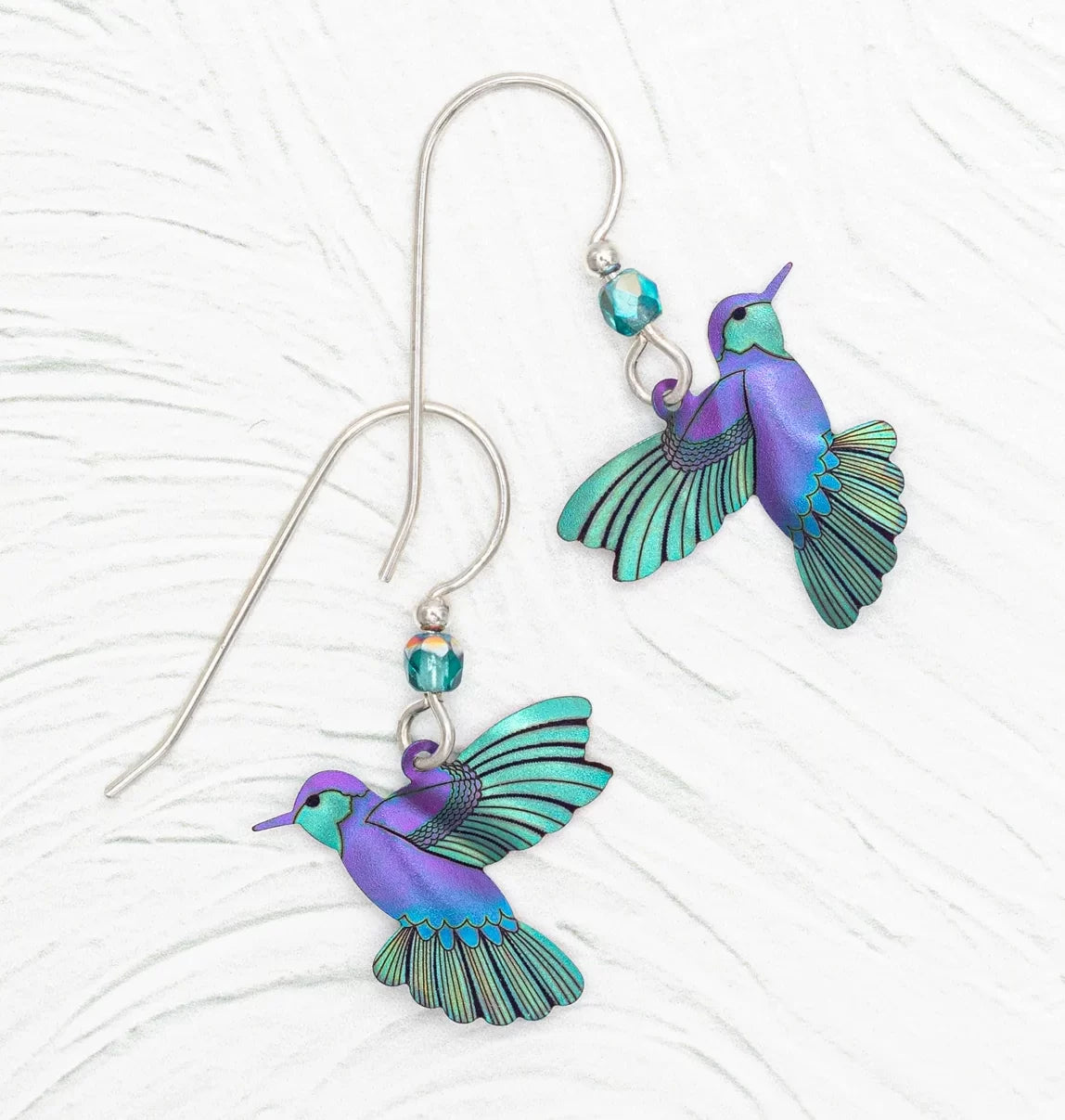 Presenting the Ultra Violet Picaflor Earrings by Holly Yashi, vibrant hummingbird-shaped treasures with purple and teal wings. Crafted from hand-painted niobium with blue faceted beads, these hypoallergenic earrings feature silver hooks against a textured white backdrop.
