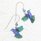 Presenting the Ultra Violet Picaflor Earrings by Holly Yashi, vibrant hummingbird-shaped treasures with purple and teal wings. Crafted from hand-painted niobium with blue faceted beads, these hypoallergenic earrings feature silver hooks against a textured white backdrop.