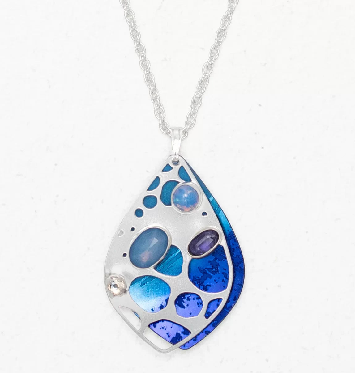 The Seashore Dreams Blue Depths Necklace by Holly Yashi features a silver teardrop pendant with blue, purple, and opalescent stones, accented by Austrian crystals. It hangs elegantly from a delicate chain against a plain white background.