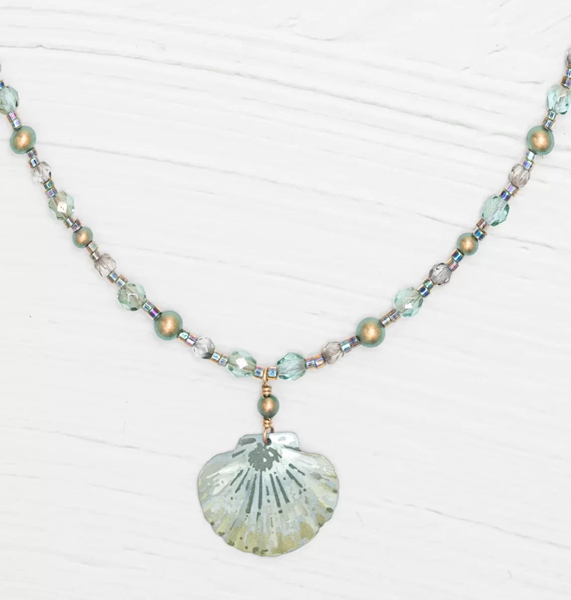The Shelby Sage and Gold Necklace by Holly Yashi features translucent and metallic beads with a seashell pendant. Crafted in light blue, silver, and light gold, this piece highlights Austrian crystals on a textured white backdrop.