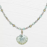 The Shelby Sage and Gold Necklace by Holly Yashi features translucent and metallic beads with a seashell pendant. Crafted in light blue, silver, and light gold, this piece highlights Austrian crystals on a textured white backdrop.