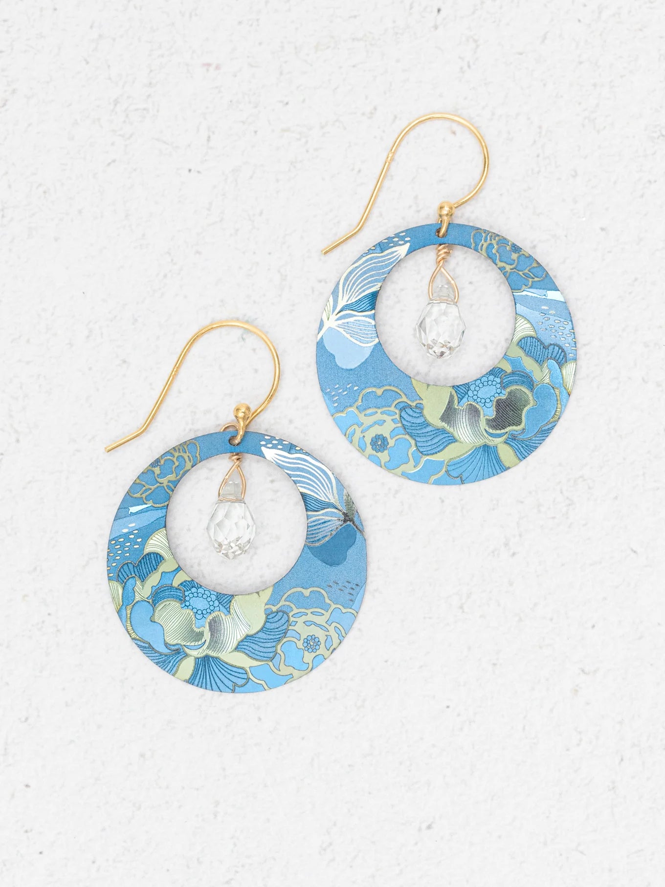 The Claire Sky Blue Earrings by Holly Yashi feature handcrafted round frames with blue, green, and gold floral patterns, gold hooks, and a clear faceted bead at the center—perfect for any occasion.