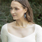 A woman with long brown hair wears a cream ribbed top, a wavy gold necklace, and Holly Yashi Turning Leaf Indigo Earrings, outdoors with greenery. She looks to her left and smiles slightly.