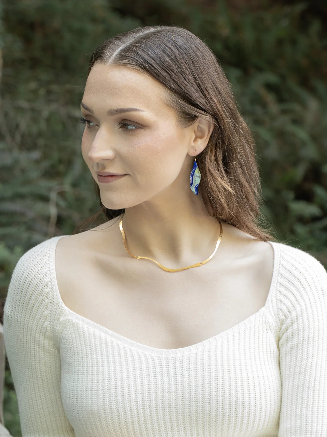 A woman with long brown hair wears a cream ribbed top, a wavy gold necklace, and Holly Yashi Turning Leaf Indigo Earrings, outdoors with greenery. She looks to her left and smiles slightly.