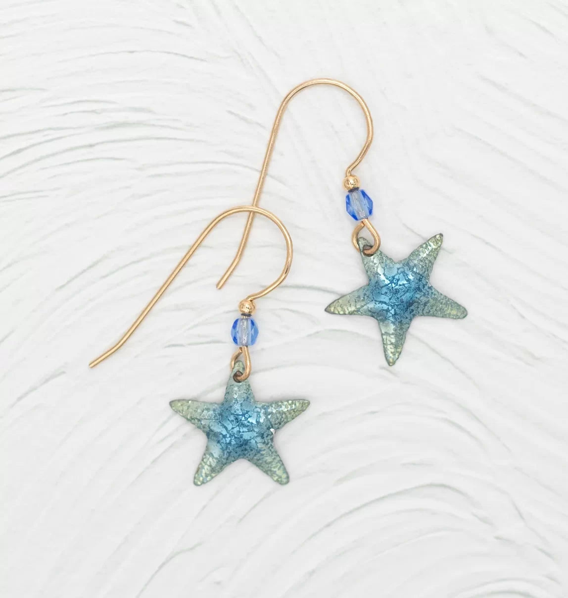 Introducing the Carmel Seashore Blue Starfish Earrings by Holly Yashi: stunning gold wire earrings featuring blue starfish pendants and small niobium beads, which are hypo-allergenic, ideal for sensitive ears. Displayed on a textured white surface.