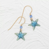 Introducing the Carmel Seashore Blue Starfish Earrings by Holly Yashi: stunning gold wire earrings featuring blue starfish pendants and small niobium beads, which are hypo-allergenic, ideal for sensitive ears. Displayed on a textured white surface.
