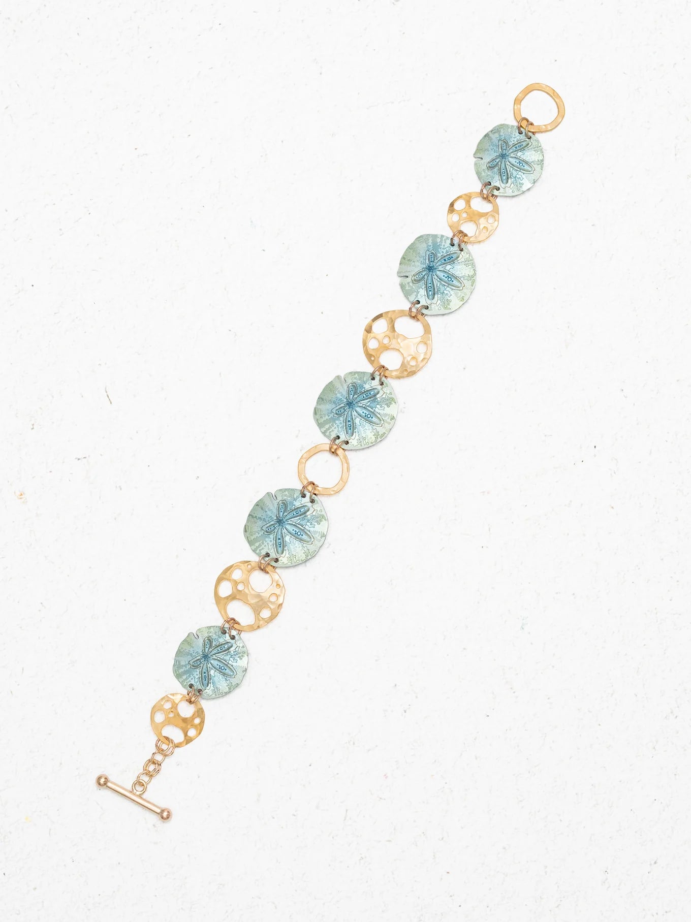 Introducing the Sage Oceana Bracelet by Holly Yashi: a stunning piece featuring alternating blue niobium links with intricate sand dollar motifs and 18k gold-plated links with elegant open circle designs, all showcased on a white speckled background.