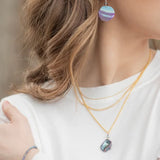 A woman with wavy brown hair wears Holly Yashi Rip Tide Calypso Earrings and layered gold necklaces featuring a dark ocean charm, complementing her white top. Her arm, adorned with a delicate hypo-allergenic bracelet, is partially visible against a blurred background.