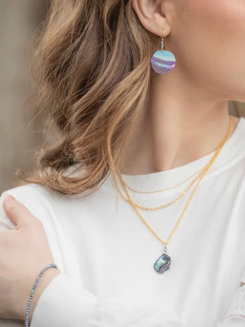 A woman with wavy brown hair wears Holly Yashi Rip Tide Calypso Earrings and layered gold necklaces featuring a dark ocean charm, complementing her white top. Her arm, adorned with a delicate hypo-allergenic bracelet, is partially visible against a blurred background.