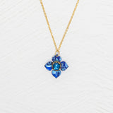 The Petite Fleur Sapphire Blue Necklace by Holly Yashi features a gold chain and a blue flower-shaped pendant with an Austrian crystal, displayed on a white textured background.