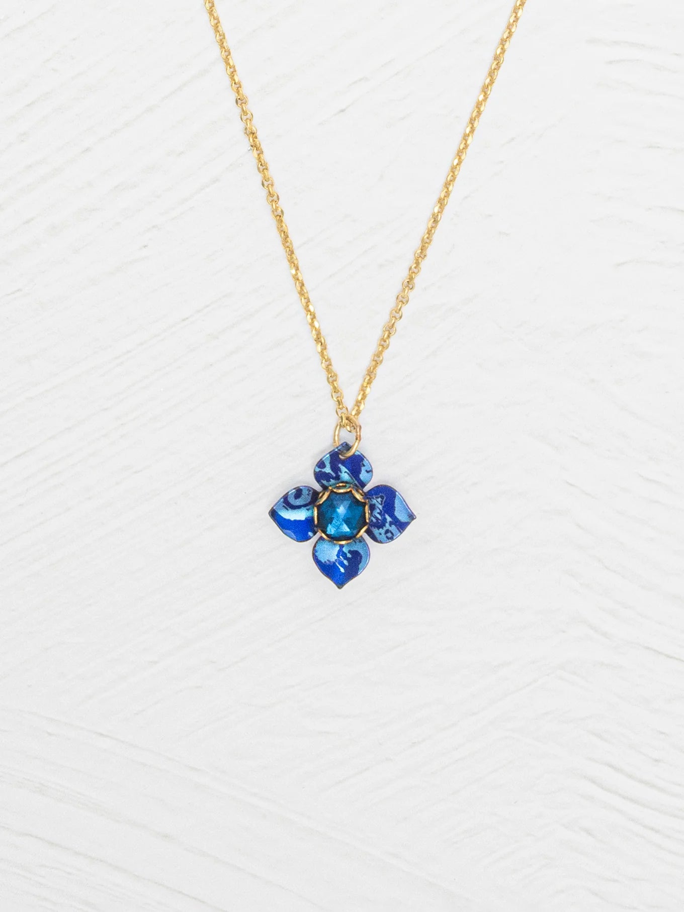 The Petite Fleur Sapphire Blue Necklace by Holly Yashi features a gold chain and a blue flower-shaped pendant with an Austrian crystal, displayed on a white textured background.