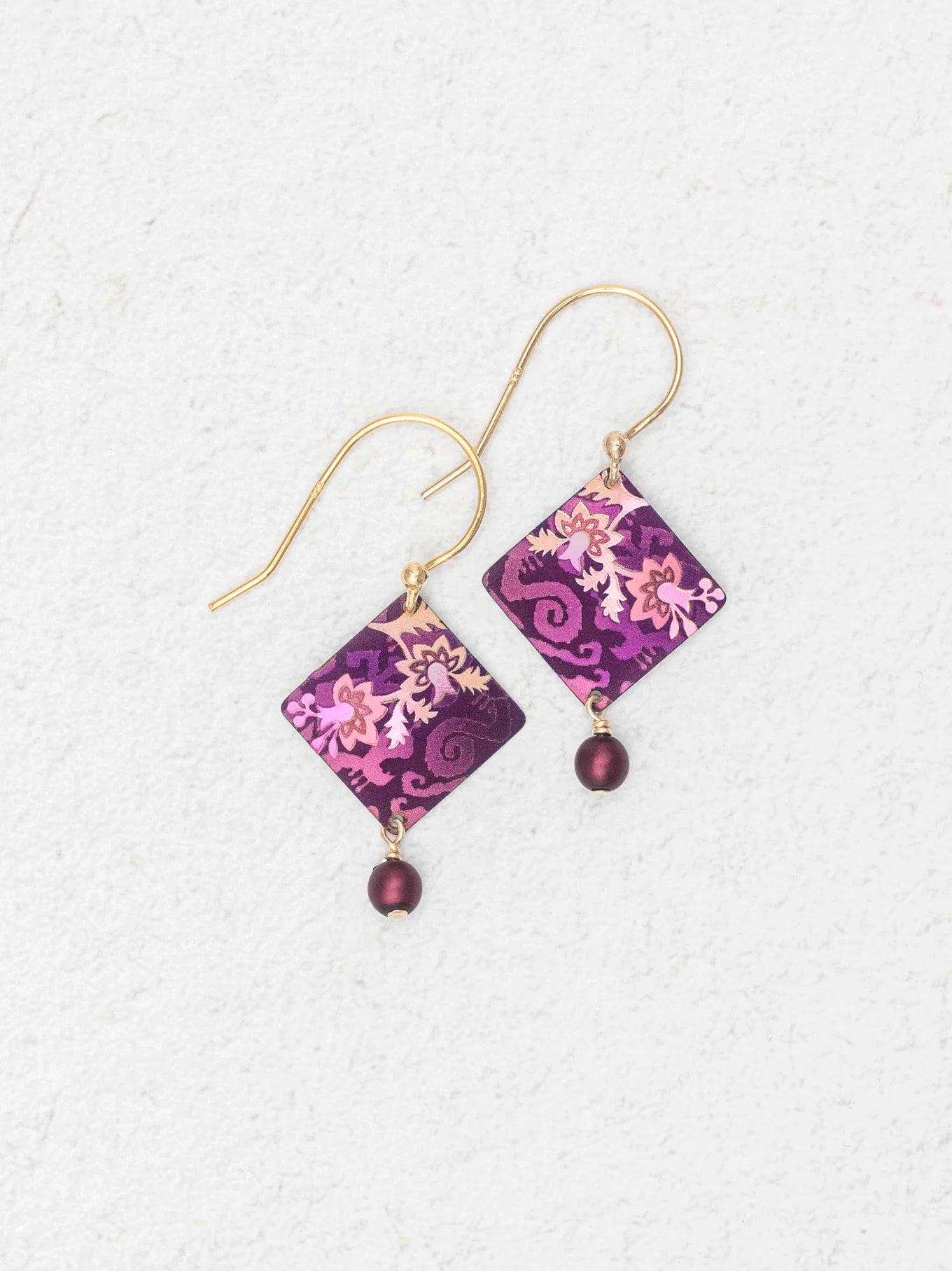 A pair of Holly Yashi Garden Soiree Vintage Burgundy Earrings, featuring gold hooks and diamond-shaped purple and pink patterned charms with a dangling round purple bead, are displayed on a light textured background.