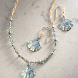 The Shelby Sage and Gold Earrings by Holly Yashi are an elegant jewelry set with a gold chain necklace, seashell earrings featuring light blue, shell-shaped pendants, and Austrian crystal pearls. The hypo-allergenic design is complemented on a light, speckled surface.