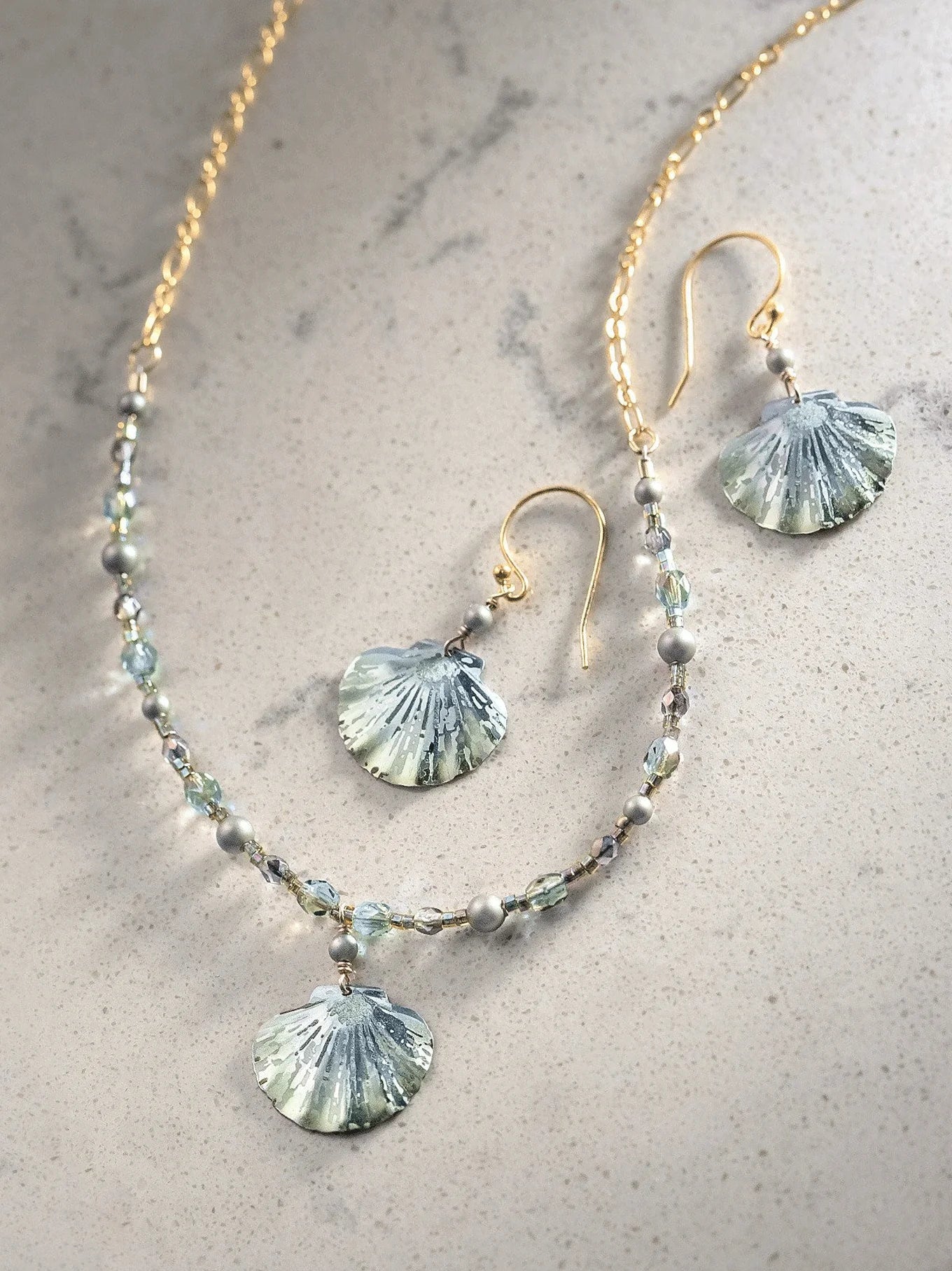 The Shelby Sage and Gold Earrings by Holly Yashi are an elegant jewelry set with a gold chain necklace, seashell earrings featuring light blue, shell-shaped pendants, and Austrian crystal pearls. The hypo-allergenic design is complemented on a light, speckled surface.