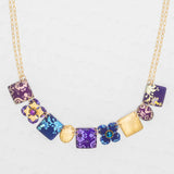 The Eliza Tiled Multi-Color Necklace by Holly Yashi features square and flower-shaped pendants in purple, blue, and gold with floral patterns and textured designs—an elegant necklace displayed on a light background.