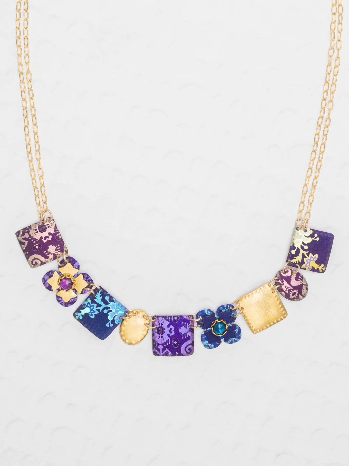 The Eliza Tiled Multi-Color Necklace by Holly Yashi features square and flower-shaped pendants in purple, blue, and gold with floral patterns and textured designs—an elegant necklace displayed on a light background.