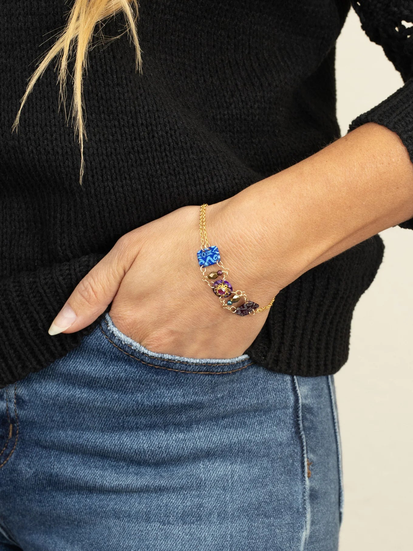 A person in a black sweater and blue jeans rests their hand in a pocket, showcasing the Holly Yashi Alex Multi Color Bracelet with vibrant, gem-inspired stones on their wrist.