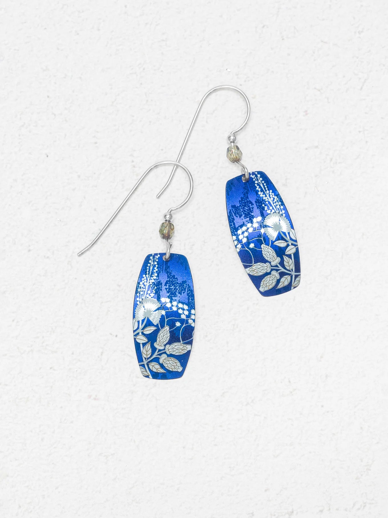 Falling Bloom Indigo Earrings by Holly Yashi: blue, oval-shaped handcrafted earrings with silver floral patterns and hooks, displayed on a light textured background.