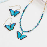 On a white surface sit the exquisite Holly Yashi Green Flash Bella Butterfly Earrings and matching necklace. The handmade set features a butterfly pendant and blue-teal beads, with each niobium earring elegantly designed as a butterfly with hooks.