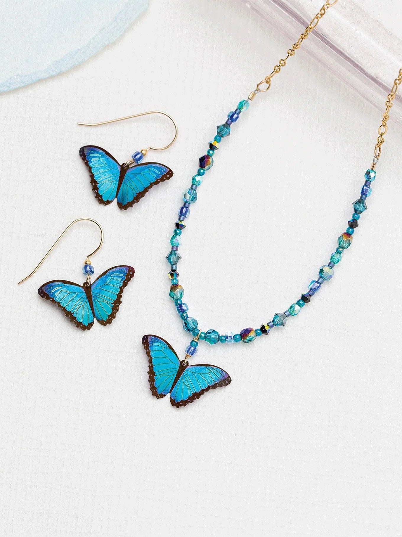 On a white surface sit the exquisite Holly Yashi Green Flash Bella Butterfly Earrings and matching necklace. The handmade set features a butterfly pendant and blue-teal beads, with each niobium earring elegantly designed as a butterfly with hooks.