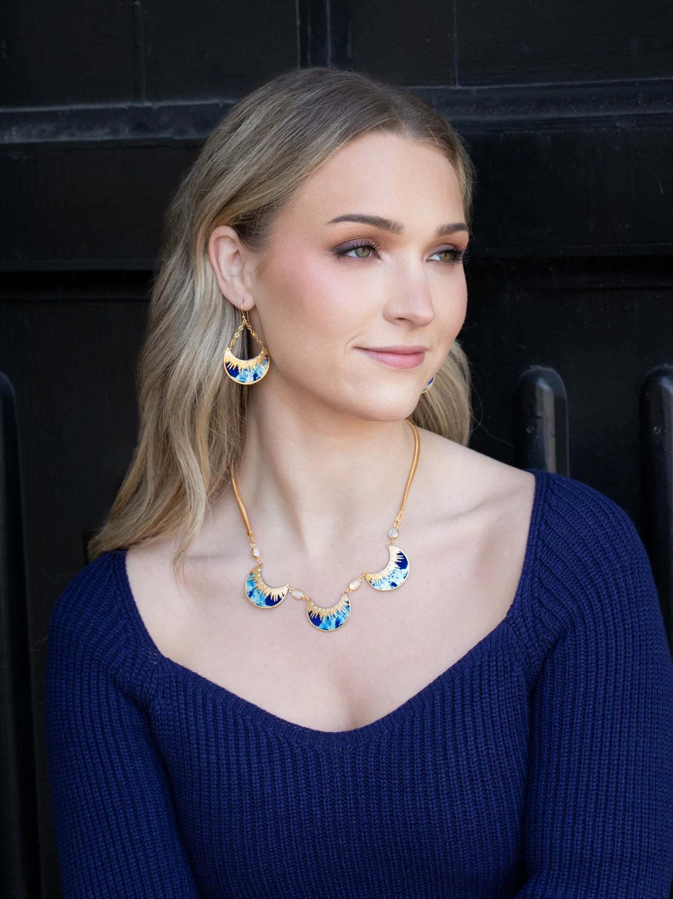 A woman with long blonde hair wears a blue knit top, colorful crescent earrings, and the Selena Necklace-Midnight Blue by Holly Yashi, sitting before a dark background and smiling softly as she looks to the side.