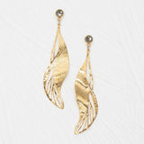 The Holly Yashi ER: Gilded Leaf Post Gold earrings feature gold, leaf-shaped drops with textured, wavy silhouettes and open cut-outs, each accented by a round crystal post. Shown against a white background.