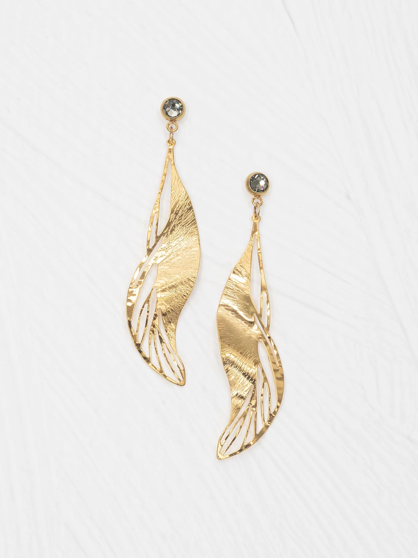 The Holly Yashi ER: Gilded Leaf Post Gold earrings feature gold, leaf-shaped drops with textured, wavy silhouettes and open cut-outs, each accented by a round crystal post. Shown against a white background.