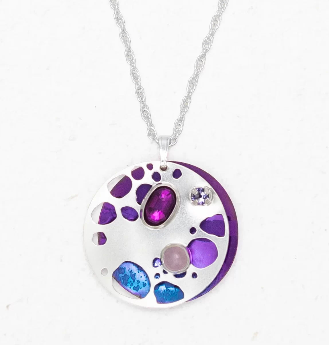 The Ocean Depths Sea Plum Necklace by Holly Yashi features a silver pendant with an abstract design, showcasing purple, blue, and pink Austrian crystals. These varied stones are set within the round pendant on a textured silver chain, offering a colorful and artistic look.