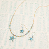 The jewelry set, placed on a textured surface, showcases a beaded necklace and Carmel Seashore Blue Starfish Earrings by Holly Yashi. Both feature star-shaped pendants with small blue beads, the hypoallergenic earrings echoing the patterned niobium bead design around the stars.