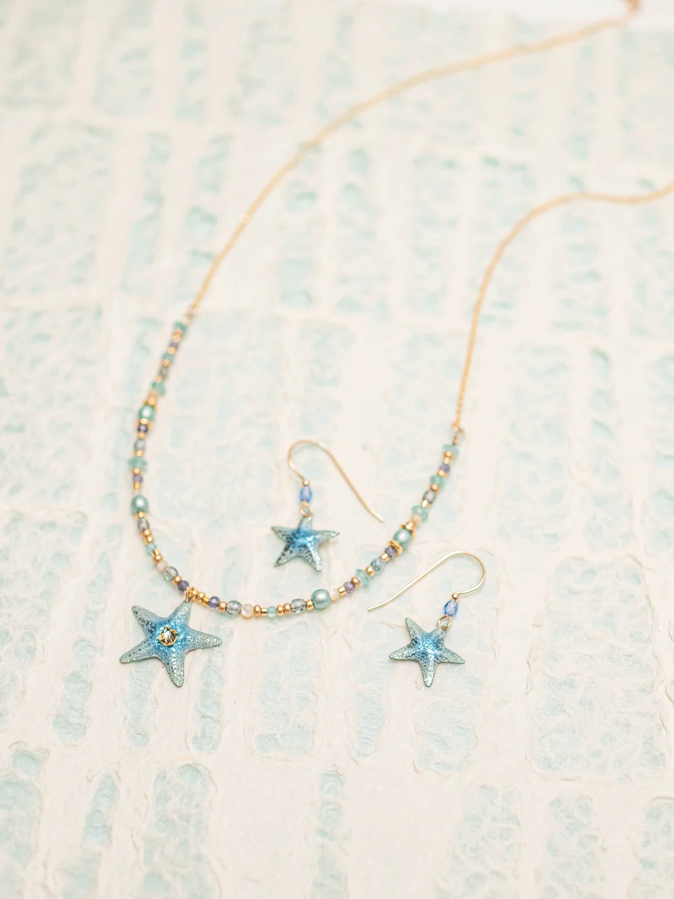 The jewelry set, placed on a textured surface, showcases a beaded necklace and Carmel Seashore Blue Starfish Earrings by Holly Yashi. Both feature star-shaped pendants with small blue beads, the hypoallergenic earrings echoing the patterned niobium bead design around the stars.