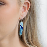 A close-up of a womans ear wearing Holly Yashis Malia Kai Blue Earrings, long oval handcrafted niobium pieces featuring a blue and light green floral pattern. Her blonde hair is tucked behind her ear, showing off the earrings.