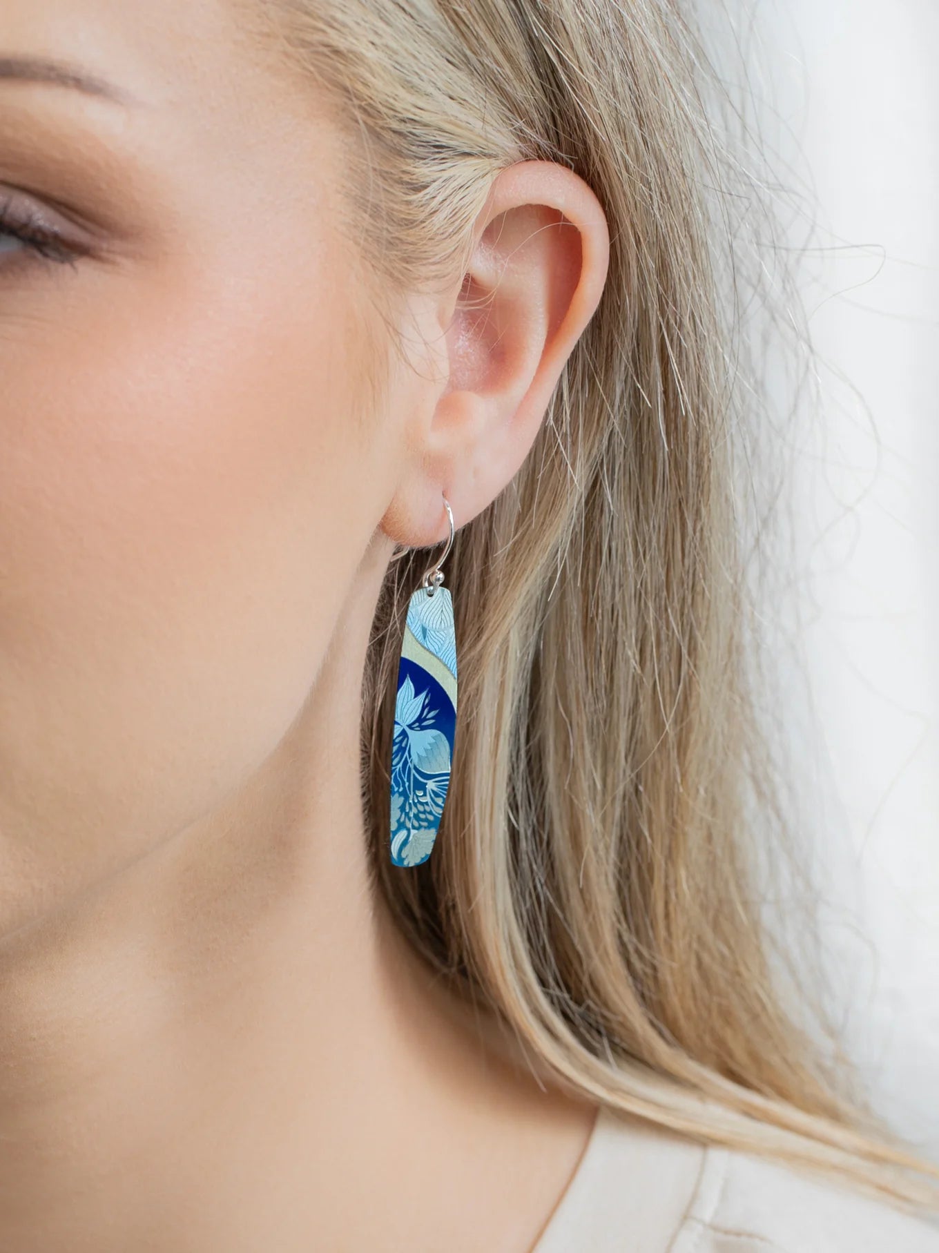 A close-up of a womans ear wearing Holly Yashis Malia Kai Blue Earrings, long oval handcrafted niobium pieces featuring a blue and light green floral pattern. Her blonde hair is tucked behind her ear, showing off the earrings.