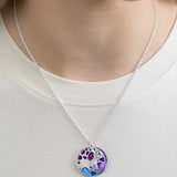 Someone styled in a light top exudes elegance while wearing the Holly Yashi Ocean Depths Sea Plum Necklace—a silver chain with a round pendant featuring Austrian crystals in vibrant purple, blue, and other shades.