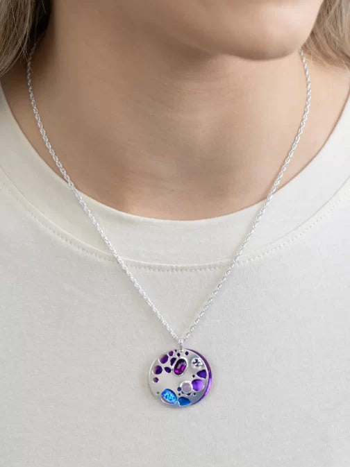Someone styled in a light top exudes elegance while wearing the Holly Yashi Ocean Depths Sea Plum Necklace—a silver chain with a round pendant featuring Austrian crystals in vibrant purple, blue, and other shades.