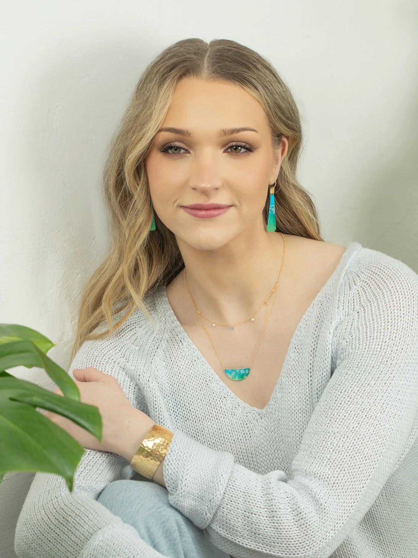A young woman with long blonde hair wears a light knit sweater, gold jewelry, and the Magnolia Turquoise/Green Necklace by Holly Yashi. She sits beside a green plant, smiling gently at the camera against a light background.