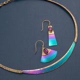 The Gianna Calypso Collar Necklace by Holly Yashi features colorful crescent-shaped accents. Its complemented by matching earrings with a vibrant gradient of teal, blue, pink, and purple stones, all hypo-allergenic and elegantly displayed on a dark background.