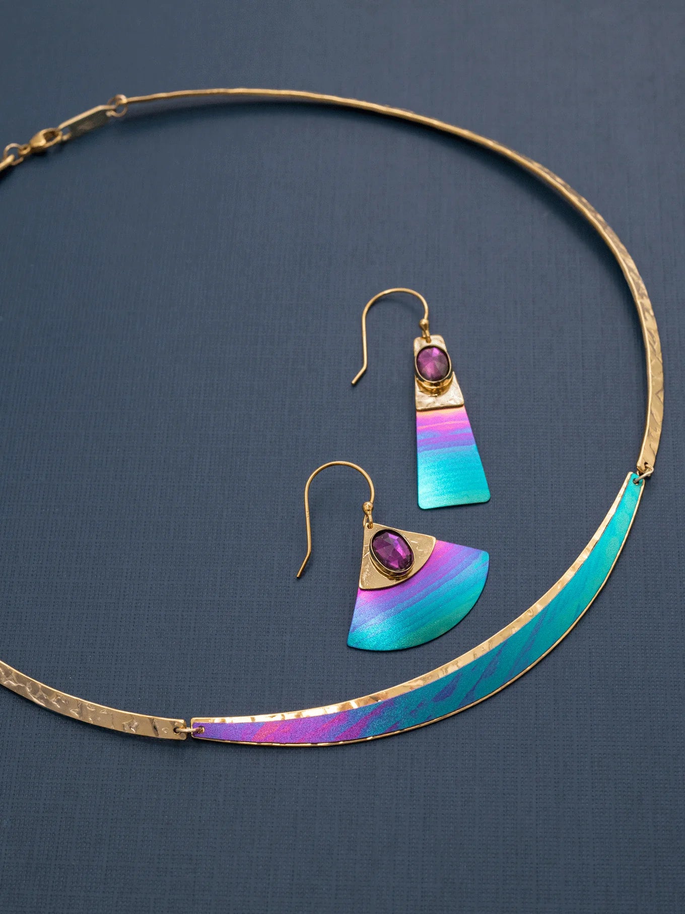 The Gianna Calypso Collar Necklace by Holly Yashi features colorful crescent-shaped accents. Its complemented by matching earrings with a vibrant gradient of teal, blue, pink, and purple stones, all hypo-allergenic and elegantly displayed on a dark background.