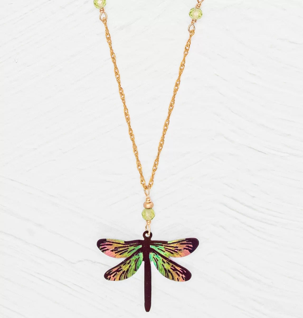 Introducing the Green Dragonfly Dreams Necklace by Holly Yashi: a gold necklace showcasing a vibrant dragonfly pendant with niobium wings in iridescent pink, green, and brown. The 18k gold plated adjustable chain is adorned with small green beads, beautifully presented on a textured white background.