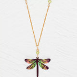Introducing the Green Dragonfly Dreams Necklace by Holly Yashi: a gold necklace showcasing a vibrant dragonfly pendant with niobium wings in iridescent pink, green, and brown. The 18k gold plated adjustable chain is adorned with small green beads, beautifully presented on a textured white background.