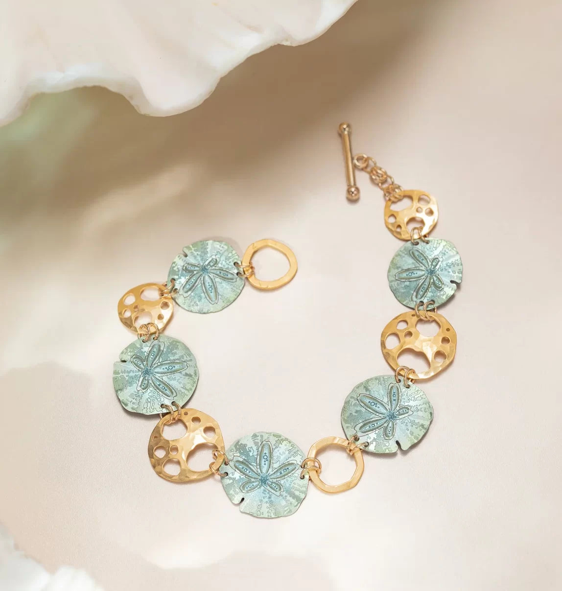 Introducing the Sage Oceana Bracelet from Holly Yashi Jewelry, featuring alternating sand dollar-like discs and 18k gold-plated circles with cutouts. A gold bar and loop clasp completes its elegance against a blurred white background.